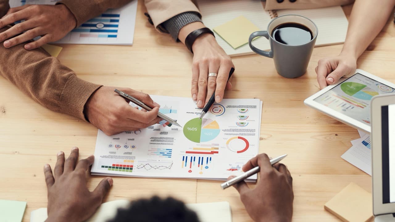 Top down view of the hands of group of people collaborating, pointing to a sheet of paper in the center of a table containing graphs and data