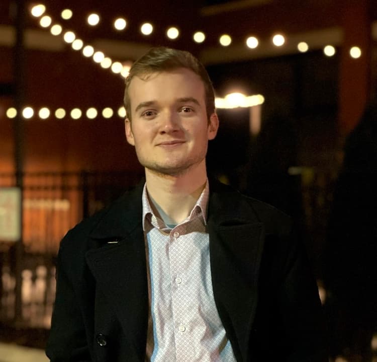 Joseph Marella, founder of Boxhouse Consulting, wearing a peacoat, collared shirt, and standing in front of some evening overhead lights